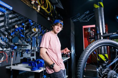 Bir genç, şık Kızıl saçlı erkek küçük işletme sahibi satış ve bisiklet tamiri mavi şapkası giyiyor ve pembe bir forma bir bisiklet dükkanda duran cep telefonu kullanıyor.