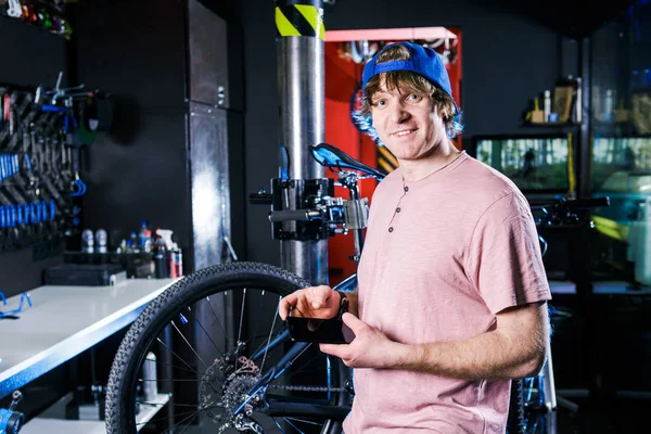 A young, stylish redheaded male small business owner selling and repairing a bike is wearing a blue cap and a pink jersey is using a mobile phone while standing in a bike shop.