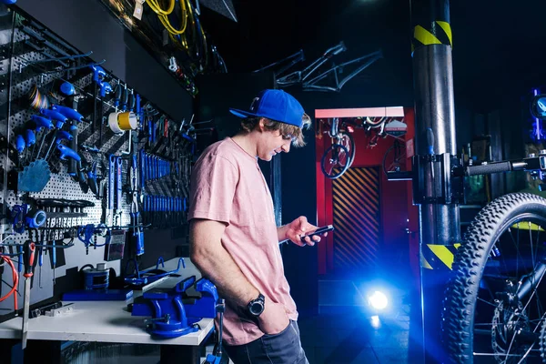 A young, stylish redheaded male small business owner selling and repairing a bike is wearing a blue cap and a pink jersey is using a mobile phone while standing in a bike shop.
