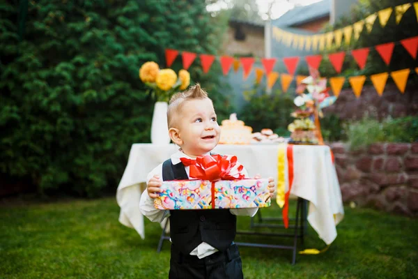 surprise for the birthday. the boy is holding a box with a gift in the yard on the background of a festive table with a cake. dressed in a black suit and a red butterfly.