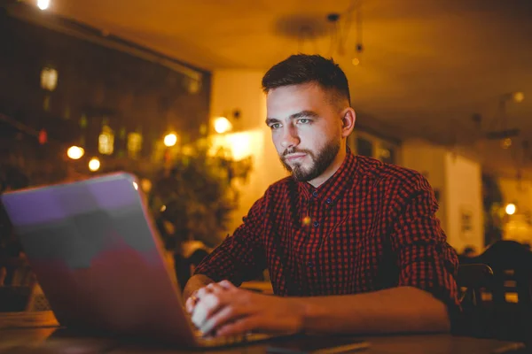 Un joven guapo hombre caucásico con barba y sonrisa dentada en una camisa roja a cuadros está trabajando detrás de un portátil gris sentado en una mesa de madera. Las manos en el teclado. Por la noche en la cafetería — Foto de Stock