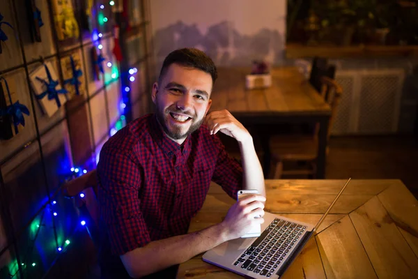 A young handsome Caucasian man with beard and toothy smile in red checkered shirt is working behind laptop sitting at wooden table. Uses holds mobile phone in hand. In the evening at the coffee shop