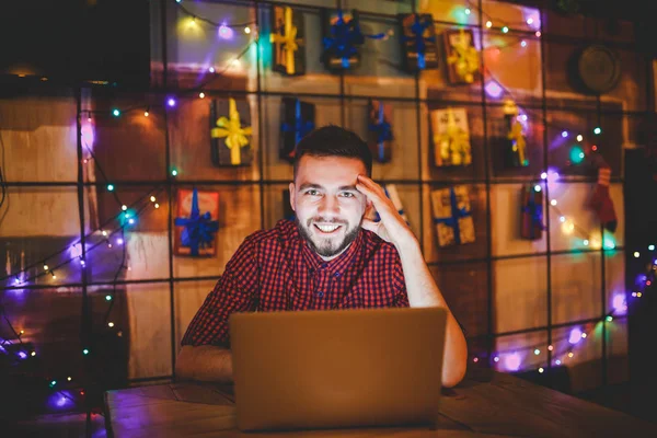 Een jonge knappe blanke man met baard en toothy glimlach in een rode geruite shirt werkt achter een grijze laptop zitten op een houten tafel. Handen op het toetsenbord. In de avond bij de coffeeshop — Stockfoto