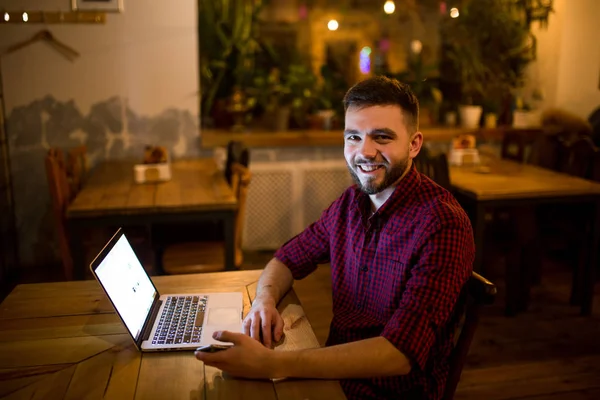 Un joven guapo hombre caucásico con barba y sonrisa dentada en camisa roja a cuadros está trabajando detrás de la computadora portátil sentada en la mesa de madera. Utiliza mantiene el teléfono móvil en la mano. Por la noche en la cafetería — Foto de Stock