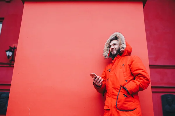 こぼれるような笑顔とひげスタンド使用、毛皮のフード付きの明るい赤冬ジャケットの赤い壁の背景にハンサムな若い男子学生は、ポケットの手で携帯電話を保持します。冬の寒さ — ストック写真