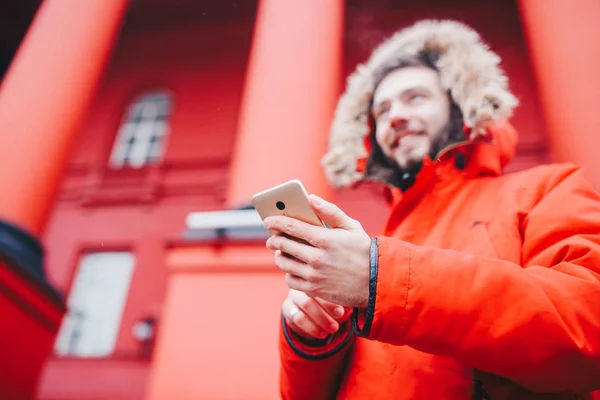 Przystojny młody mężczyzna student z Uśmiech zębiczny i broda stoi na tle czerwone ściany, fasady instytucji edukacyjnych w Czerwona zimowa kurtka kaptur z futerkiem, używa palec na ekranie telefonu komórkowego — Zdjęcie stockowe