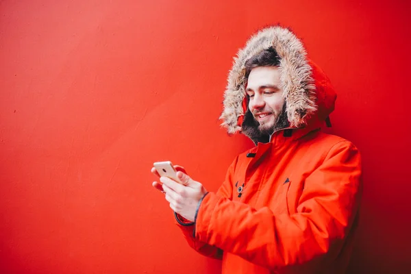 Handsome young male student with toothy smile and beard stands on red wall background, facade of educational institution in red winter jacket with hood with fur, Uses finger on screen of mobile phone — Stock Photo, Image
