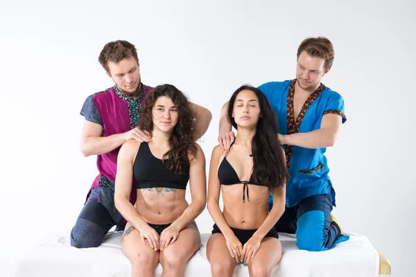 Theme massage and body care. Two male Caucasian twin brothers in uniform doing shoulder massage to two beautiful sexy European women in lingerie sitting on massage table ga isolated white background.
