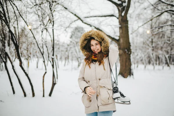 美しい若い白人女性フードやファーはジーンズで彼女の頭の上にジャケットで雪に覆われた公園に立って 彼女の肩に長い靴ひもの白スケートのペアを保持しています — ストック写真