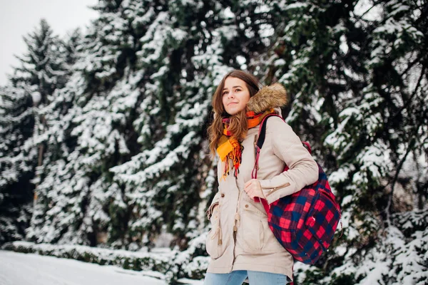 Theme is weekend holiday in winter. A beautiful young Caucasian woman stands in snow park in jacket with hood and fur in jeans and smiles with checkered backpack on background of New Year tree.