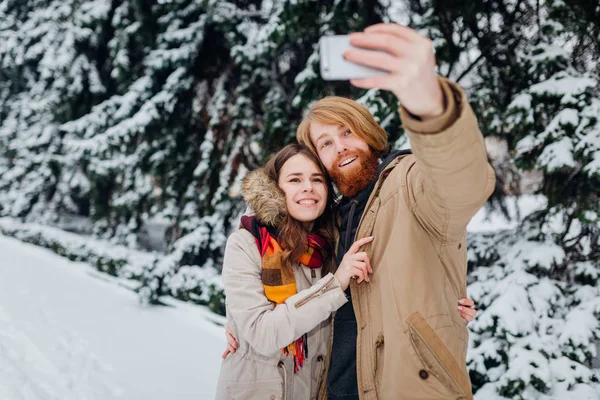 Young couple in love make joint photo of themselves on mobile phone. The guy holds smartphone on his elongated hand and makes selfie photo on background of park with coniferous trees in winter.