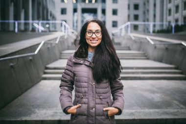 Güzel ve genç birbayan dişlek bir gülümsemeyle uzun esmer saçlı Avrupa etnik köken, gözlük ve bir ceket giyen bir iş merkezi fonunda bulutlu bulutlu hava sonbaharda duruyor.