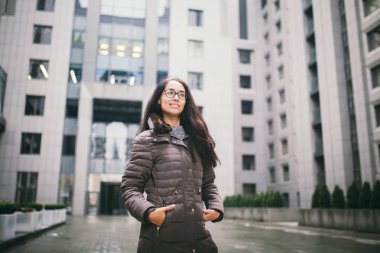 Güzel ve genç birbayan dişlek bir gülümsemeyle uzun esmer saçlı Avrupa etnik köken, gözlük ve bir ceket giyen bir iş merkezi fonunda bulutlu bulutlu hava sonbaharda duruyor.