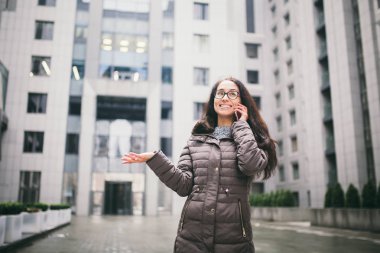 İş durum temasıdır. Avrupa elbise giyen gözlük ve kat uzun esmer saçlı güzel genç kadın iş merkezi arka plan üzerinde duruyor ve telefon elinde yakın kullanır.