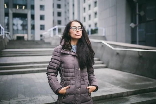 Beautiful young woman of European ethnicity with long brunette hair, wearing glasses and a coat stands against backdrop of a business center in the fall in cloudy cloudy weather.