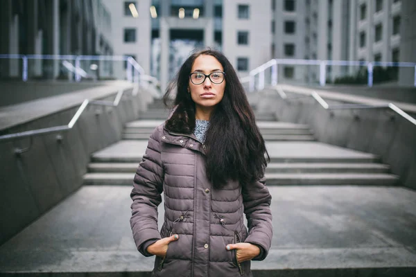 Beautiful young woman of European ethnicity with long brunette hair, wearing glasses and a coat stands against backdrop of a business center in the fall in cloudy cloudy weather.