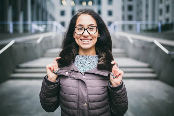 Beautiful young woman of European ethnicity with long brunette hair with a toothy smile, wearing glasses and a coat stands against backdrop of a business center in the fall in cloudy cloudy weather.