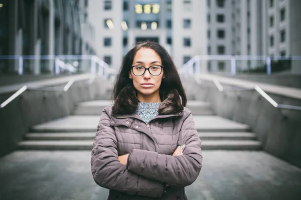 Beautiful young woman of European ethnicity with long brunette hair with grave emotion, wearing glasses and a coat stands against the backdrop of a business center in fall in cloudy cloudy weather.