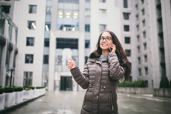 Theme is the business situation. Beautiful young woman of European ethnicity with long brunette hair wearing glasses and coat stands on background of business center and uses phone in hand near ear.