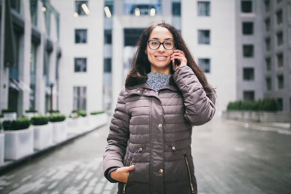 Theme is the business situation. Beautiful young woman of European ethnicity with long brunette hair wearing glasses and coat stands on background of business center and uses phone in hand near ear.