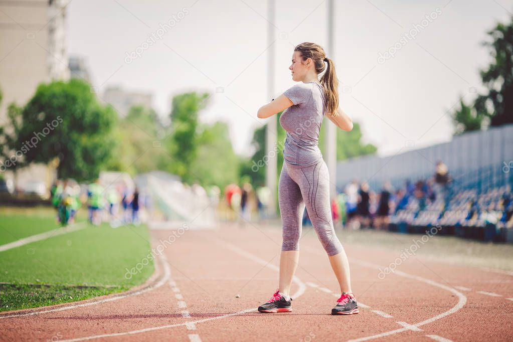 Beautiful young caucasian woman with long hair in tail and big breasts doing exercises, warming up and warming up muscles before training in running stadium, red rubber track in summer on a sunny day.