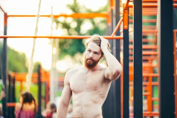 Subject sport street workout Handsome young caucasian man with a bare-chested muscular with long red hair and beard posing outdoor sports ground, gym outdoors. Sexy guy touches his hair