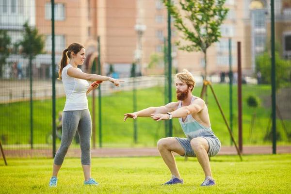 Team work in sports. female trainer training man with beard in park on green grass, lawn at sunset. The girl holds phone in hand and uses stopwatch, guy does exercises for the buttocks of sitting down