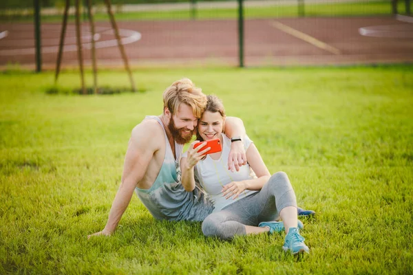 Sport et technologie. Jeune amoureux hétérosexuel couple caucasien se reposant après l'entraînement à l'extérieur dans le parc sur la pelouse, herbe verte assis dans l'étreinte et faire selfie photo sur smartphone caméra au coucher du soleil — Photo