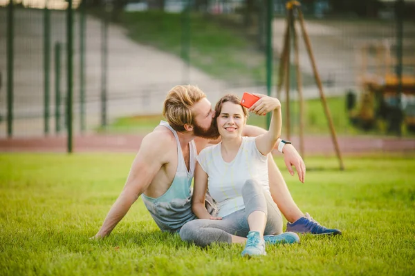 Desporto e tecnologia. Jovens apaixonados heterossexuais casal caucasiano descansando após o treino ao ar livre no parque no gramado, grama verde sentado no abraço e fazendo foto selfie na câmera do smartphone ao pôr do sol — Fotografia de Stock