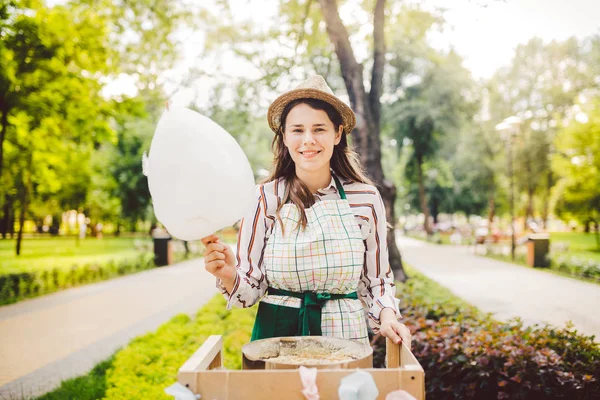 Фото теми малого бізнесу приготування солодощів. Молода біла жінка з продавцем фартухів у капелюсі власник розетки робить цукерки муліне, казкову муліне або бавовняну цукерку в літньому парку — стокове фото