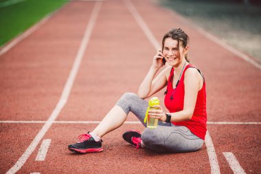 Spor ve teknoloji. At kuyruğu oturma Stadyumu'nda, kırmızı koşu bandı parça çalışma sırasında egzersiz yaptıktan sonra dinlenme ile bir güzel genç beyaz bir kadın. Cep telefonu kulaklık ve spor saatler kullanımları.
