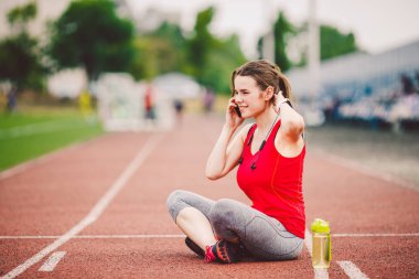 Spor ve teknoloji. At kuyruğu oturma Stadyumu'nda, kırmızı koşu bandı parça çalışma sırasında egzersiz yaptıktan sonra dinlenme ile bir güzel genç beyaz bir kadın. Cep telefonu kulaklık ve spor saatler kullanımları.