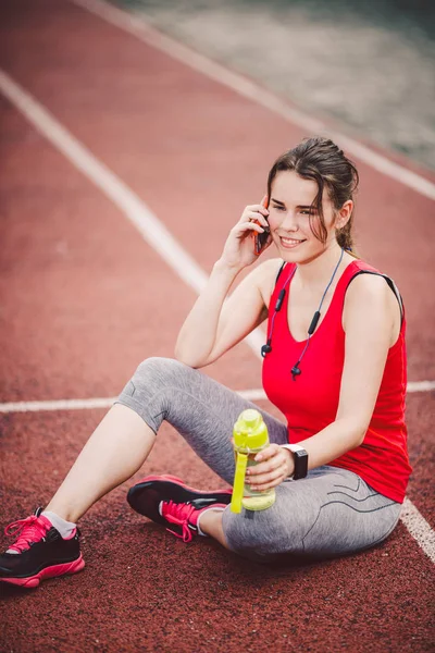 Sport and technology. A beautiful young Caucasian woman with ponytail sitting resting after workout during run at stadium, a red treadmill track. Uses for cell phone headphones and sports watches.