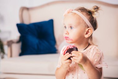 Çocukların doğum günü. İki yaşında, pembe elbiseli komik bir beyaz kız. Evin yatak odasında duruyor. Kanepenin arkasında yemek yiyor, elinde pasta tatlısı tutuyor. Yüzü ve ağzı krem bulaşmış..