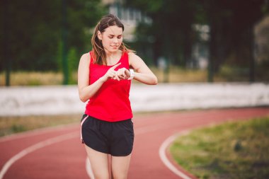 Güzel genç atlet beyaz kırmızı T-shirt ve jogging, koşu stadyumda kısa şort büyük meme ile bir kadın. At kuyruğu saç modeli bir spor pulseometer basın için eline bir parmak kullanır