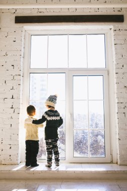Tema Noel sabahı. İki bebek beyaz erkek ve kız kardeşi ve kız kardeşi kucaklama büyük kutusunda pencere önü sırtında ile kışın ayakta güneşli bir günde Tuhafiye yün elbise ve şapka evin.