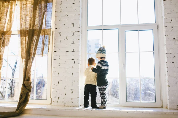 Theme Christmas morning. Two baby Caucasian boy and girl brother and sister standing with backs on window front of large box in embrace in winter knitted woolen clothes and hat of house in sunny day.