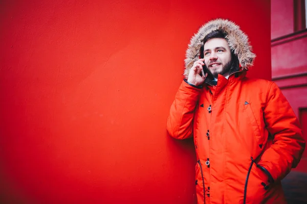 こぼれるような笑顔とひげのハンサムな若い男子学生が冬の毛皮フード付きの明るい赤冬ジャケットの赤い壁の背景に立っています 携帯電話 トークや通話を使用します — ストック写真