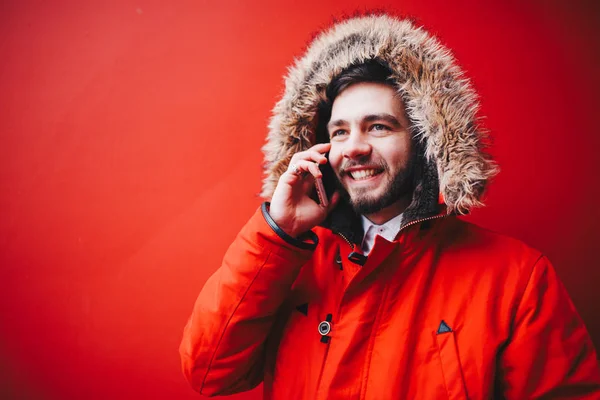 こぼれるような笑顔とひげのハンサムな若い男子学生が冬の毛皮フード付きの明るい赤冬ジャケットの赤い壁の背景に立っています 携帯電話 トークや通話を使用します — ストック写真