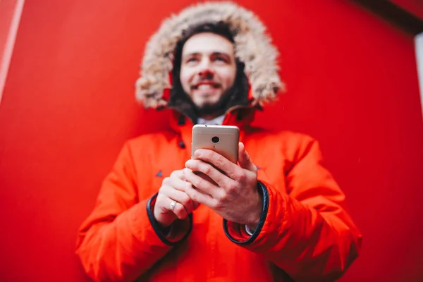 こぼれるような笑顔とひげのハンサムな若い男子学生が赤い壁背景 毛皮のフード 携帯電話の画面で使用する指と赤いジャケットの教育機関のファサードの上に立つ — ストック写真