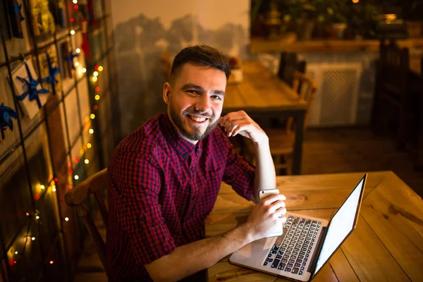 A young handsome Caucasian man with beard and toothy smile in red checkered shirt is working behind laptop sitting at wooden table. Uses holds mobile phone in hand. In the evening at the coffee shop.