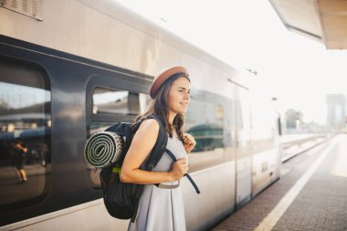 Tema demiryolu ve seyahat. Dişlek ile genç beyaz kadın portre gülümseme, sırt çantası ve Turizm izole mat elbise ve şapka yaz aylarında araçlar ile Tren İstasyonu Tren arka plan ayakta.