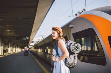 Tema demiryolu ve seyahat. Dişlek ile genç beyaz kadın portre gülümseme, sırt çantası ve Turizm izole mat elbise ve şapka yaz aylarında araçlar ile Tren İstasyonu Tren arka plan ayakta.