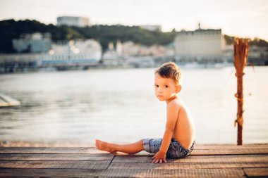 Bir çocuk ve yaz plaj tatil temasıdır. Küçük bir beyaz çocuk yana doğru bir ahşap iskele üzerinde oturur ve kumlu bir plaj ve bir su birikintisi bir nehir kameraya bakar. Çıplak ayaklı mavi kot şort.