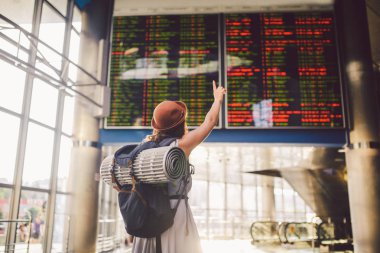 Tema seyahat toplu taşıma. İçeri elbise ve şapka arkasında duran genç kadın sırt çantası uyku için kamp ekipmanları, yalıtım mat zamanlama çetele Havaalanı İstasyonu elinde cepheye görünüyor.