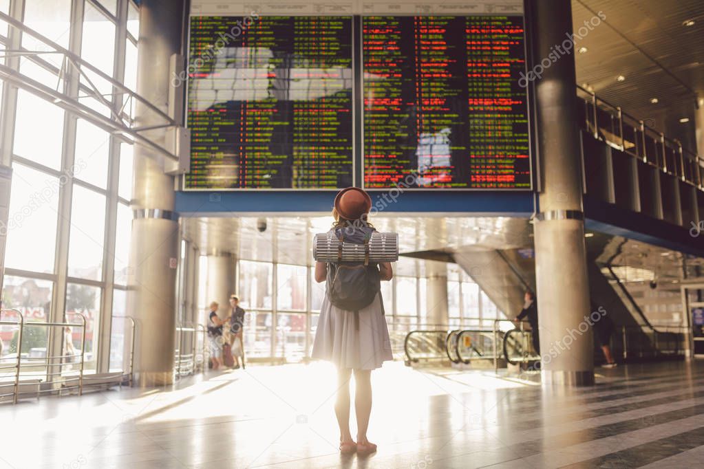 Theme travel public transport. young woman standing with back in dress and hat behind backpack and camping equipment for sleeping, insulating mat looks schedule on scoreboard airport station sunny day