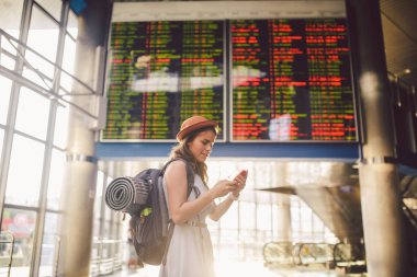 Tema seyahat ve tranosport. Güzel genç kafkas kadın elbise ve tren istasyonuna veya kırmızı telefon tutarak bir zamanlama seyir terminal içinde duran sırt çantası iletişim teknolojisini kullanır