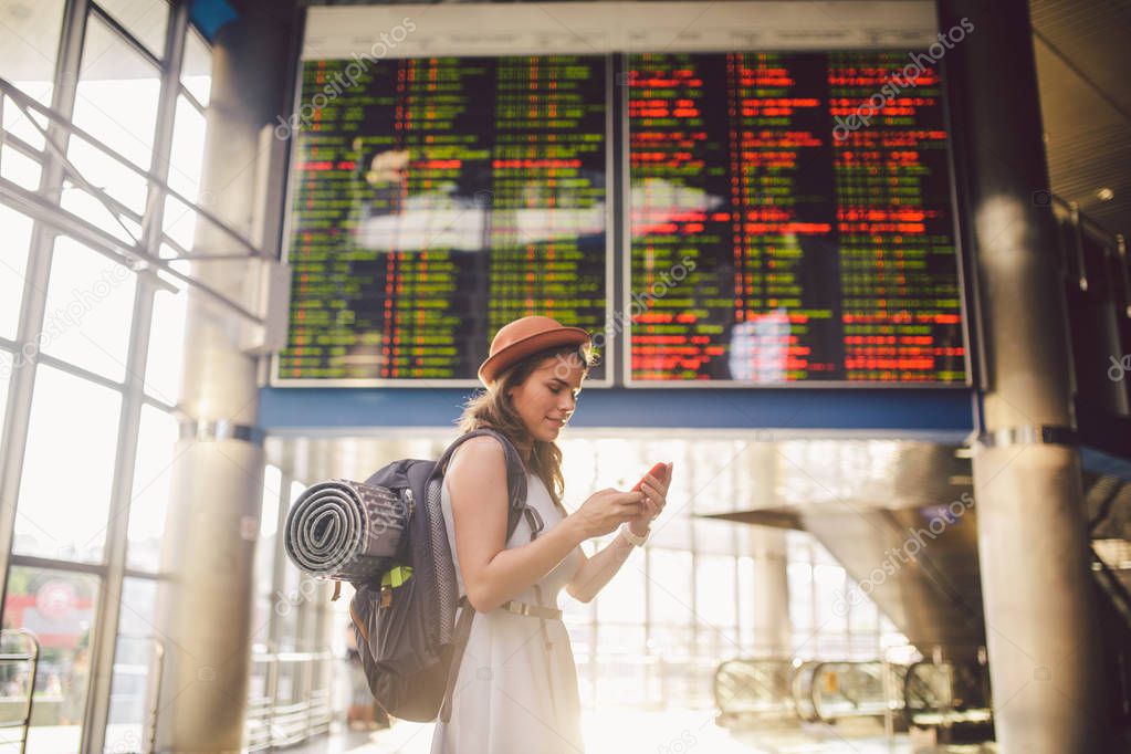 Theme travel and tranosport. Beautiful young caucasian woman in dress and backpack standing inside train station or terminal looking at a schedule holding a red phone, uses communication technology