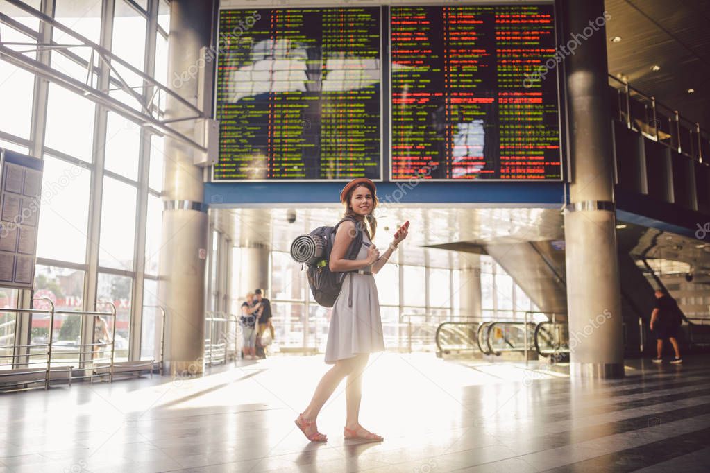 Theme travel and tranosport. Beautiful young caucasian woman in dress and backpack standing inside train station or terminal looking at a schedule holding a red phone, uses communication technology