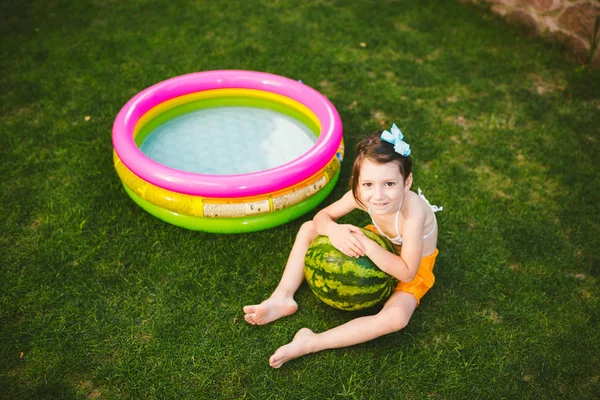 El tema de la alimentación adecuada niños sanos. Niña 4 años caucásica sentada sobre hierba verde abrazando grandes bayas redondas verdes sandía cerca de los niños piscina inflable en el patio de la casa — Foto de Stock